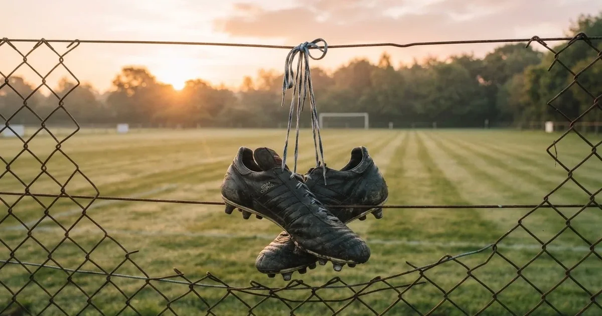 Znoszone buty piłkarskie zawieszone za sznurówki na drucianym płocie na skraju boiska treningowego o wschodzie słońca.
