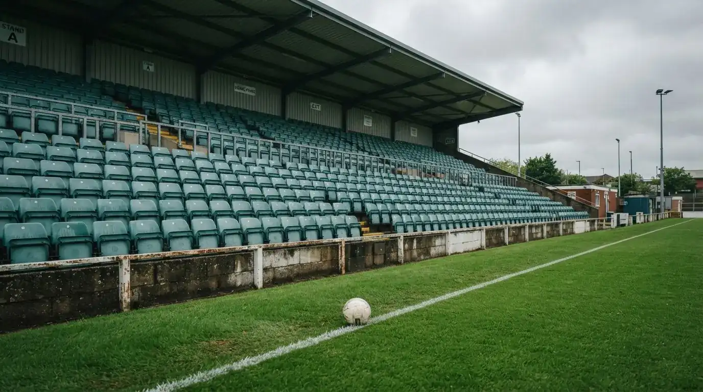 Rzędy pustych plastikowych krzesełek małego stadionu widziane z poziomu murawy pod pochmurnym niebem.