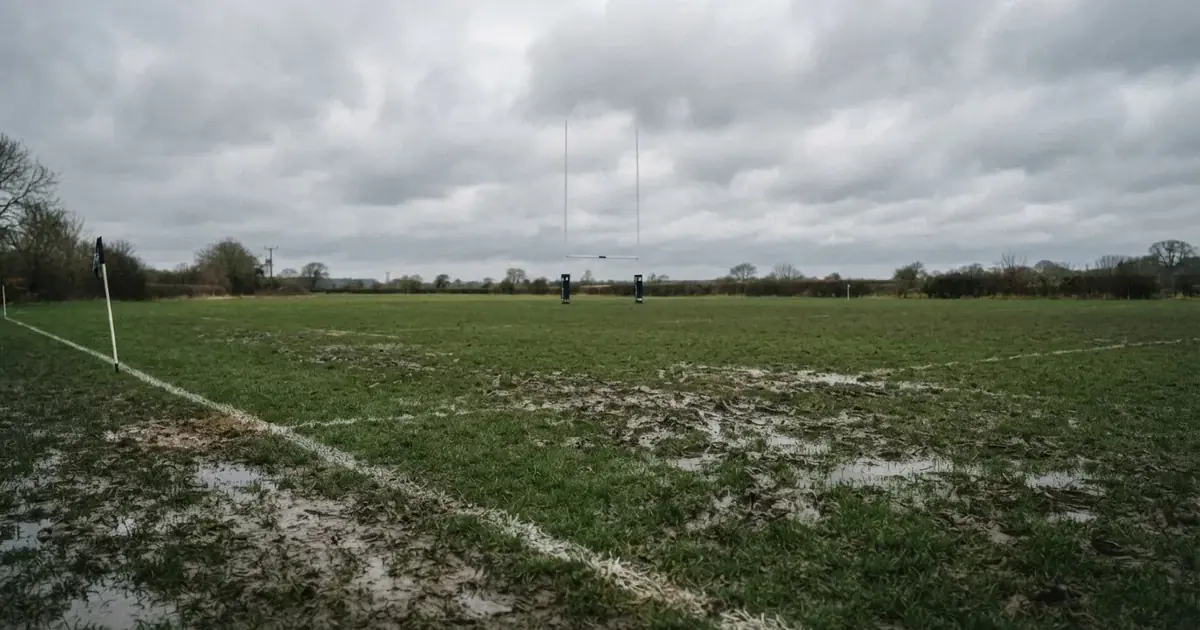 Boisko do rugby ze słupami bramki w oddali pod pochmurnym niebem, mokra trawa i ślady butów.